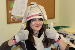 A smiling woman in a knit hat, scarf and mittens giving a thumbs-up in an office