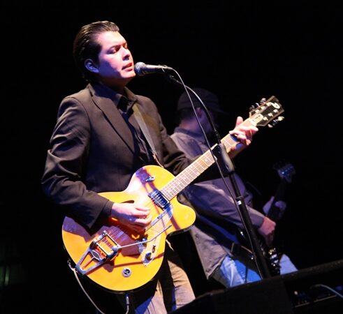 A man in a dark jacket singing into a microphone and playing a yellow hollow-body guitar on a dark stage
