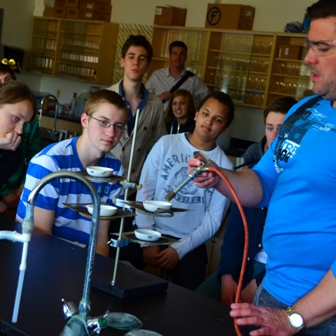 Students gather around an instructor demonstrating a Bunsen burner experiment in a chemistry lab