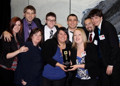 Nine smiling people in dark clothing crowd together with their arms around each other, holding a black award trophy