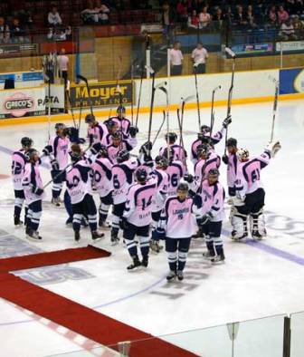A women's hockey team in pink and navy jerseys raises their sticks in salute on the ice at a Pink in the Rink game