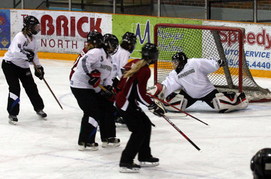 Women's hockey players in white and maroon fight for the puck in front of the net, as a goalie in white sprawls on the ice