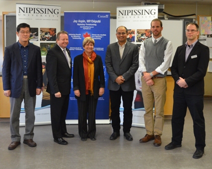 Six people stand in a row in a classroom in front of Nipissing University banners, including one for Jay Aspin, MP
