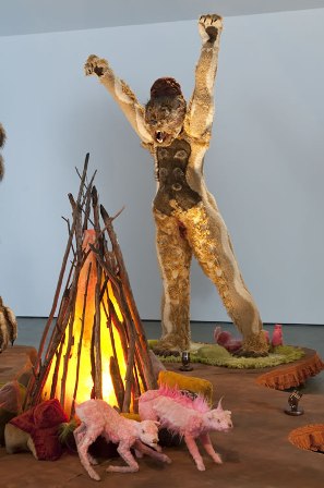 A sculpture of a figure with raised arms standing behind a glowing teepee-shaped fire and two pink animal figures in a gallery