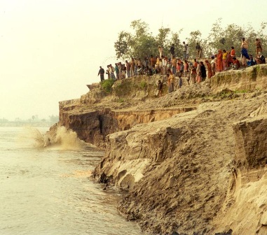 A crowd of people standing on top of a crumbling riverbank cliff above rushing brown water
