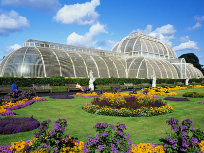 A large glass greenhouse with a formal garden of purple and yellow flowers in front