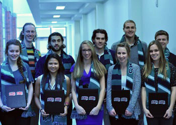 Ten students wearing striped scarves and holding black folders pose together in a hallway