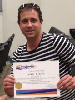 A smiling man in a striped shirt holding a certificate in his name for a best overall concurrent session award at a conference