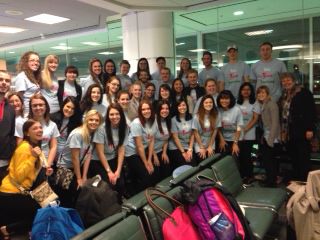 A large group of young women in matching light grey t-shirts posing together in an airport waiting area