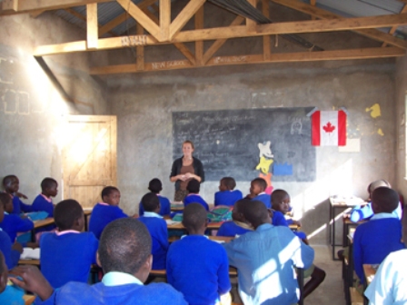 A woman teaches a classroom of children in blue uniforms in a simple building with a Canadian flag on the wall
