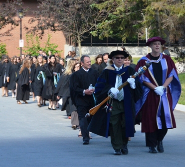 Faculty in academic robes carrying a ceremonial mace lead a procession of graduates in black gowns down a path