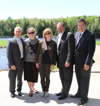 Five people stand together on a paved area beside a lake with trees behind them