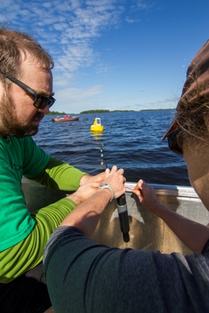 Two researchers on a boat lowering a probe into a lake, with a yellow buoy in the distance