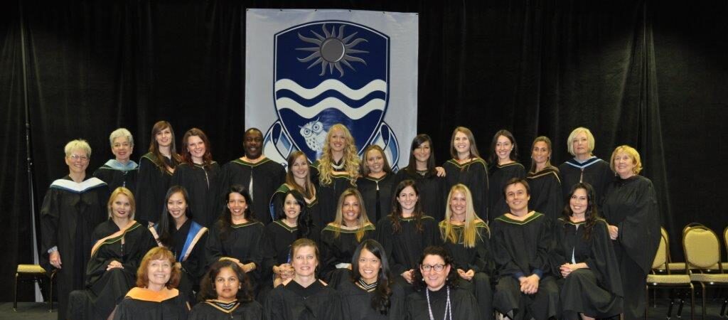 A large group of women in graduation gowns seated and standing on a stage in front of a Nipissing University banner