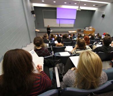View from the back of a lecture hall of students seated at desks, facing an instructor standing at the front