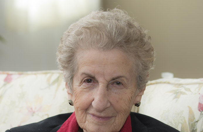 Portrait of an older woman with short grey hair, wearing a black jacket and red blouse, seated on a floral sofa