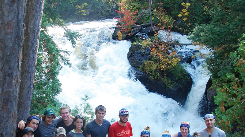 A large group of students in athletic clothes and headbands posing on rocks above a rushing waterfall