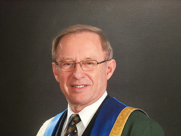 Portrait of a smiling man in glasses wearing a green, blue and gold academic robe