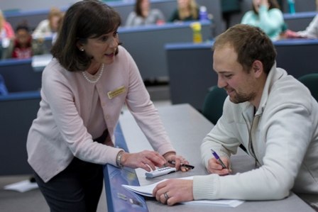 A woman in a pink cardigan leaning over a lecture-hall desk to help a man write in a notebook