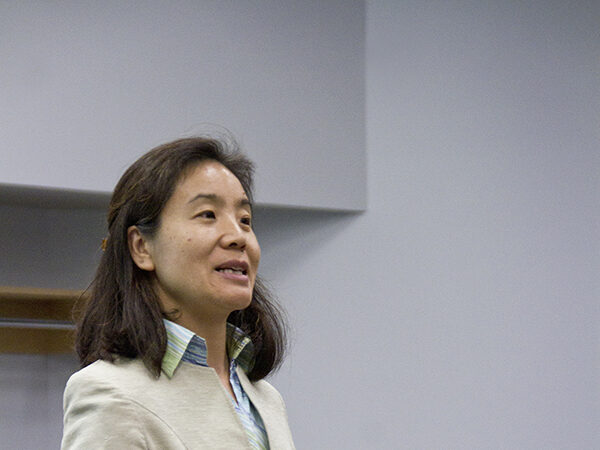 A woman in a beige jacket gesturing as she speaks to a classroom, with a student seated behind her at a laptop