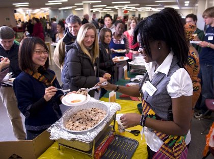 People lining up to be served food from a buffet by a woman in a vest and African print apron at a large indoor event