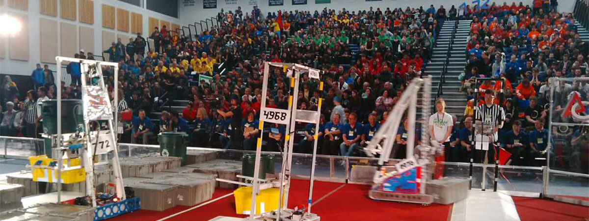 A robotics competition arena with a red carpeted field and robots, with a large crowd in the stands