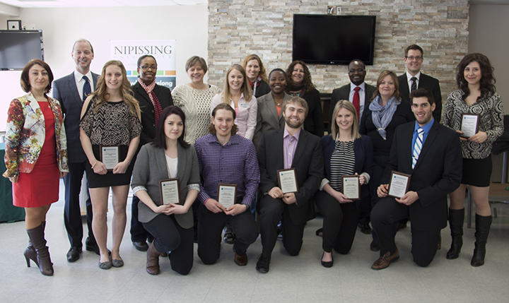 A group of about twenty students and staff pose in rows in front of a stone fireplace wall, several holding award plaques