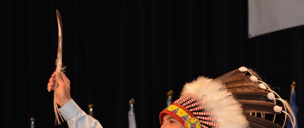 A man in a feathered headdress holding up an eagle feather on stage in front of flags