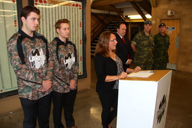 Two Nipissing hockey players in camouflage jerseys stand beside a woman signing a book at a podium, with two soldiers and a man in a suit behind