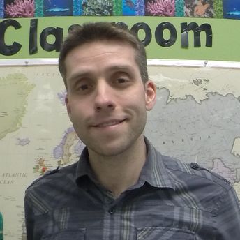 A man with short brown hair smiling at the camera in front of a world map with the word 'Classroom' behind him
