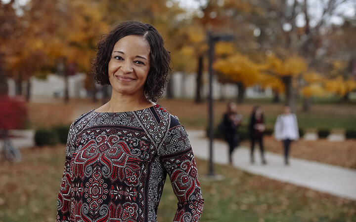 A smiling woman with curly hair in a patterned top standing on a campus path in autumn