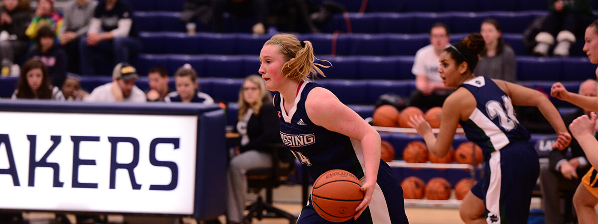 A Nipissing women's basketball player in navy dribbles the ball down the court as opponents follow