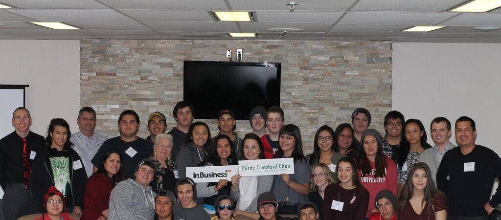 A large group of students and staff posing for a photo indoors, holding signs that read 'InBusiness Purdy Crawford Chair in Aboriginal Business Students' and 'Cape Breton University Happen'
