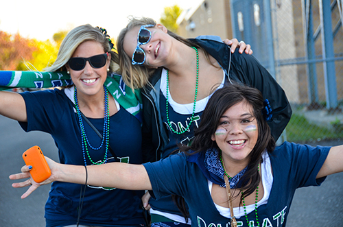 Three laughing young women in navy Nipissing shirts, face paint and green beads posing with outstretched arms