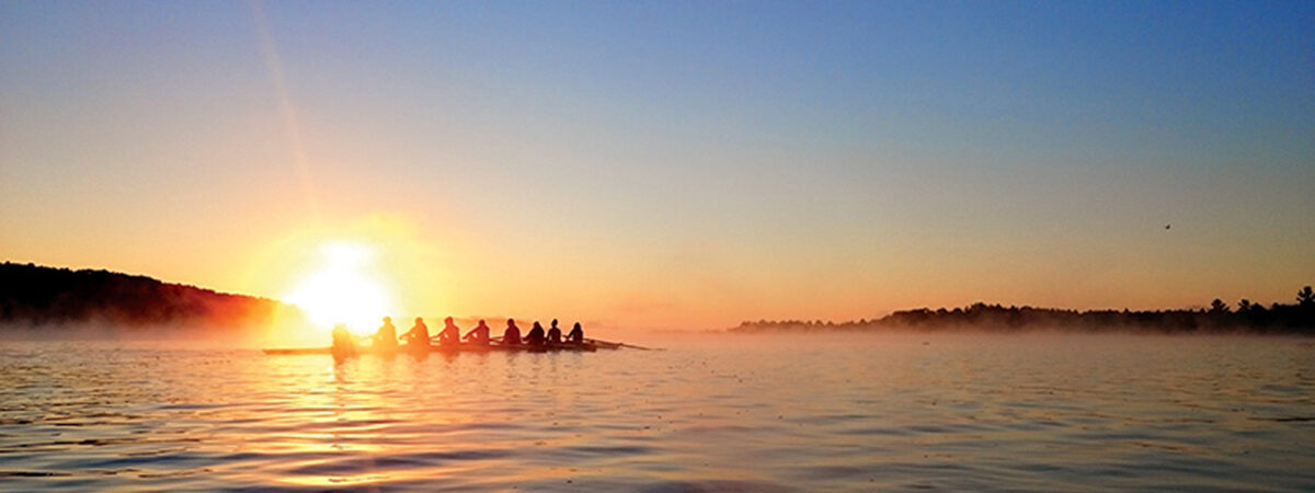 A rowing crew paddling across a misty lake at sunrise, silhouetted against the rising sun