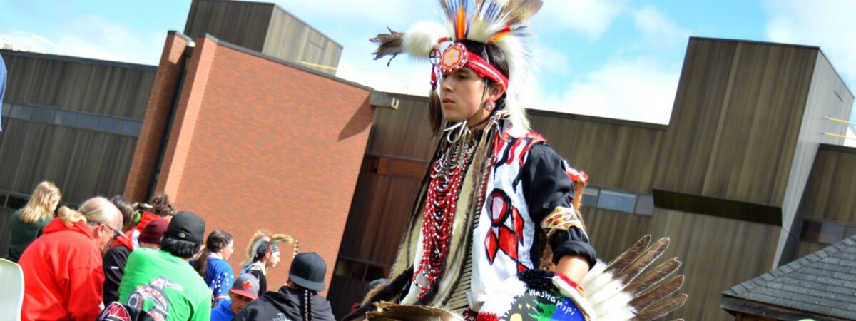 A young man in a feathered powwow regalia dancing on a lawn while an audience sits on chairs behind him