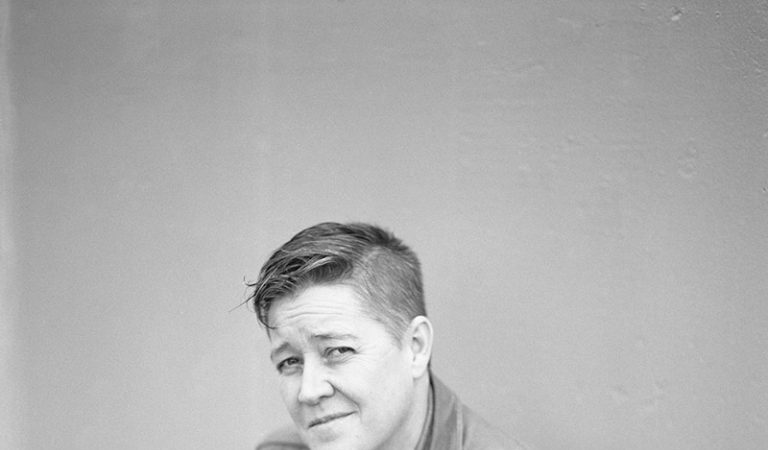 Black and white portrait of a person with short hair, in a denim shirt, crouching and looking at the camera