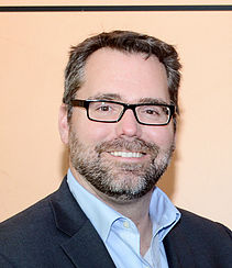 Portrait of a smiling man with glasses and a beard, wearing a navy blazer and light blue shirt