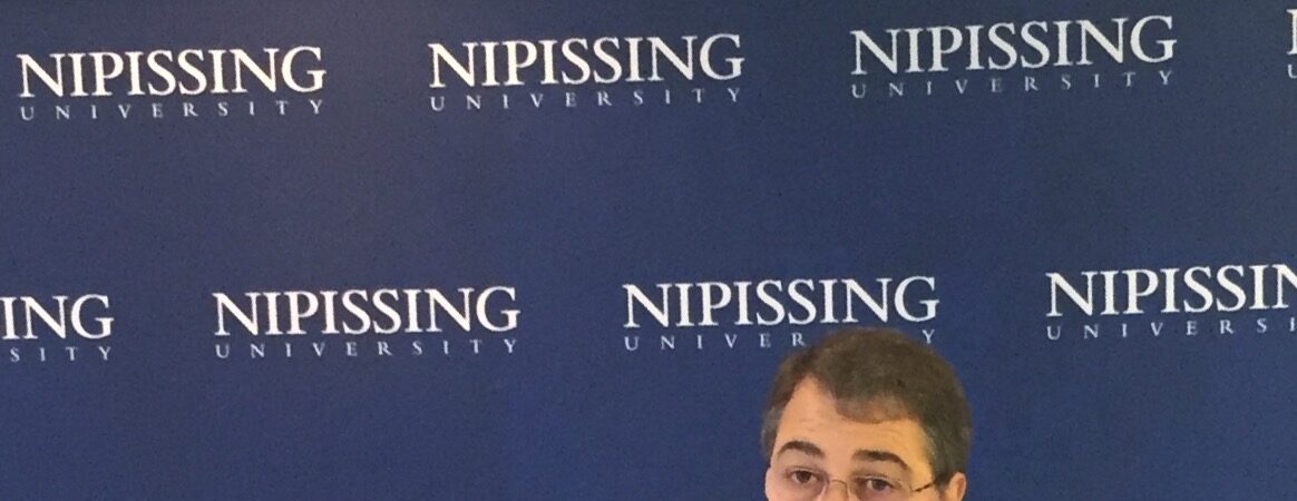 A man in a dark suit speaking from a Nipissing University podium in front of a Nipissing University backdrop