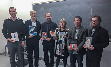 Six people stand in a row in front of a chalkboard, each holding up a book