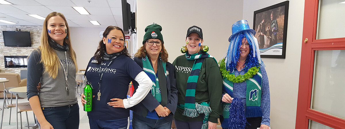 Five smiling staff members dressed in Nipissing green and blue clothing, scarves and wigs, posing in a hallway
