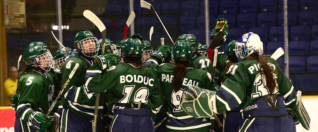 A women's hockey team in green and navy jerseys huddles with sticks raised on the ice