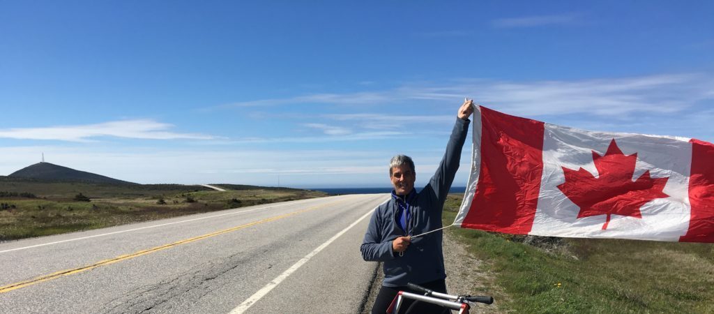 A man holding up a Canadian flag beside a road, standing next to a shopping cart with signs on it
