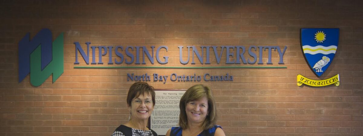 Two women shaking hands and holding an envelope in front of a Nipissing University sign with the university crest