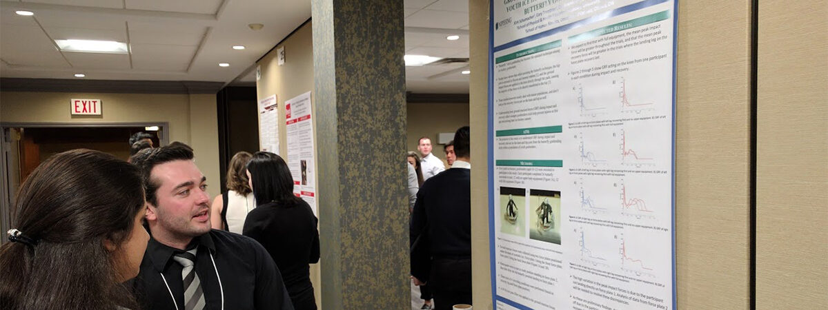 A man in a dark suit and lanyard chatting with a woman beside a scientific research poster in a conference hallway