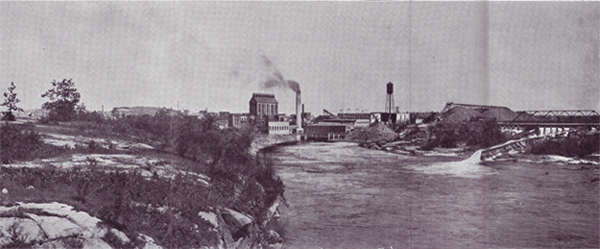 Black and white historical photo of a mill and a river with rapids, with smokestacks and a water tower in the distance