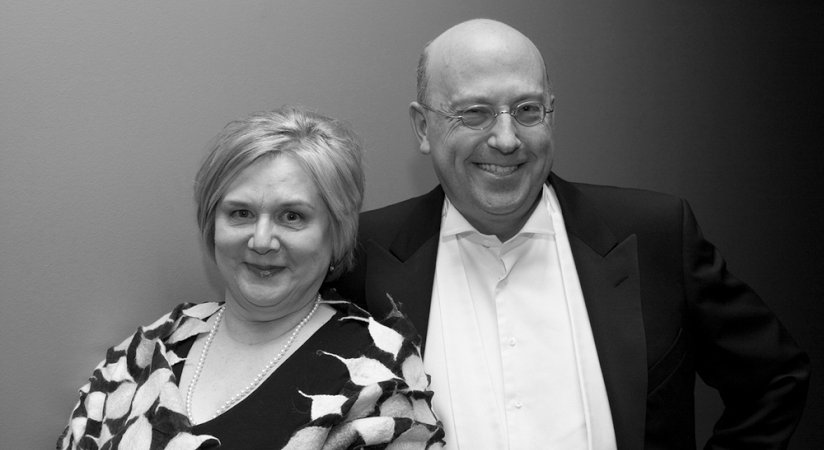 Black and white portrait of a smiling couple, a woman in a patterned jacket and a bald man in a black tuxedo and glasses