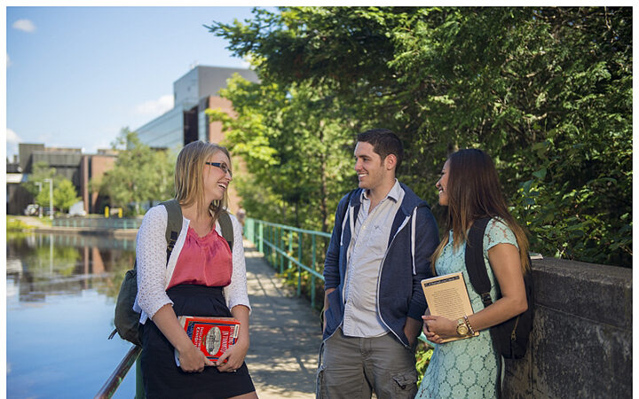 Three students chatting and smiling on a path beside a pond, carrying books, with campus buildings behind them