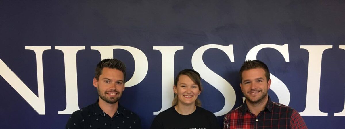 Two men and a woman stand in front of a large Nipissing wall sign; the woman wears a Nipissing shirt and the men hold a certificate and a box of cupcakes