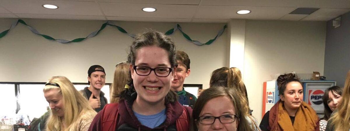 Two smiling students with their arms around each other in a crowded lounge, one holding a slice of pizza